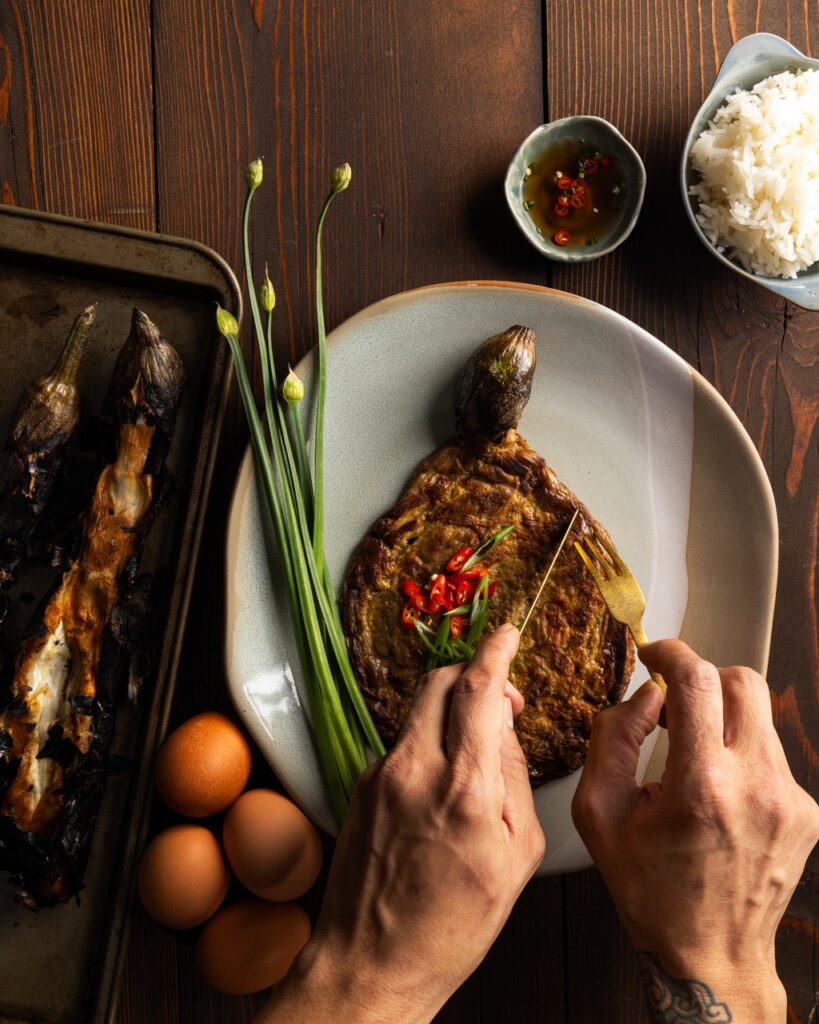 A person uses a fork and knife to cut into an eggplant omelet on a white plate with chives and roasted egg plants sitting beside it.