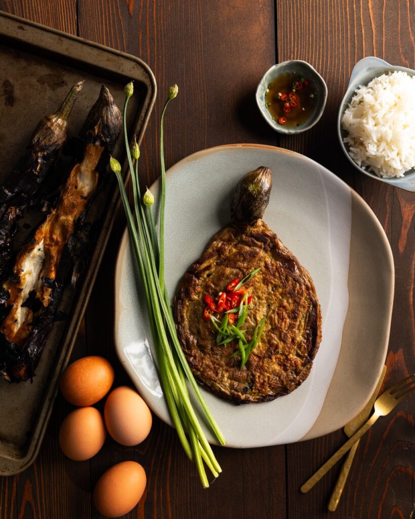 A round white plate with a round Tortang Talong Eggplant Omelet in the center with chives beside it and eggplants on a baking sheet nearby.