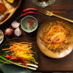julienned vegetables sit on a green plate beside other ingredients and a brown plate with a large Filipino fritter with shrimp on top of it.