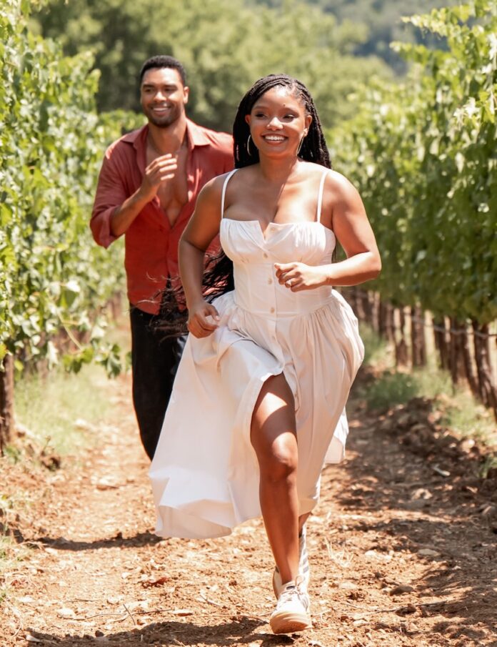 Halle Bailey runs through a vineyard in a white dress in Tuscany with Regé-Jean Page behind her.