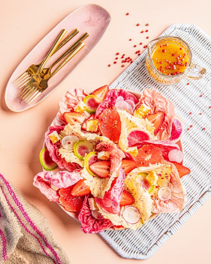 A bowl full of pink radichio and strawberries with a pink peppercorn vinaigrette in the upper right corner and gold silverware in the left.