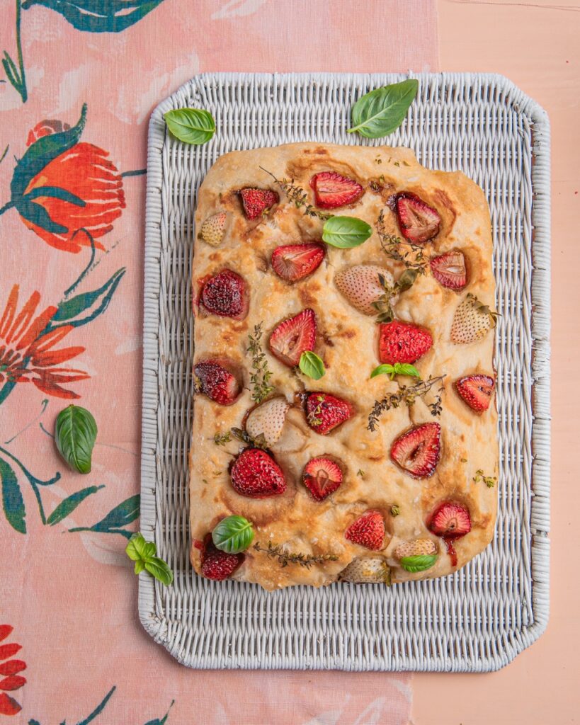 A loaf of Strawberry Focaccia with basil leaves on top of a wire platform on top of a pink floral table.