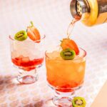 A cocktail pours from a cocktail shaker into a glass to make a Strawberry Kiwi Bramble with kiwi and strawberry garnish.