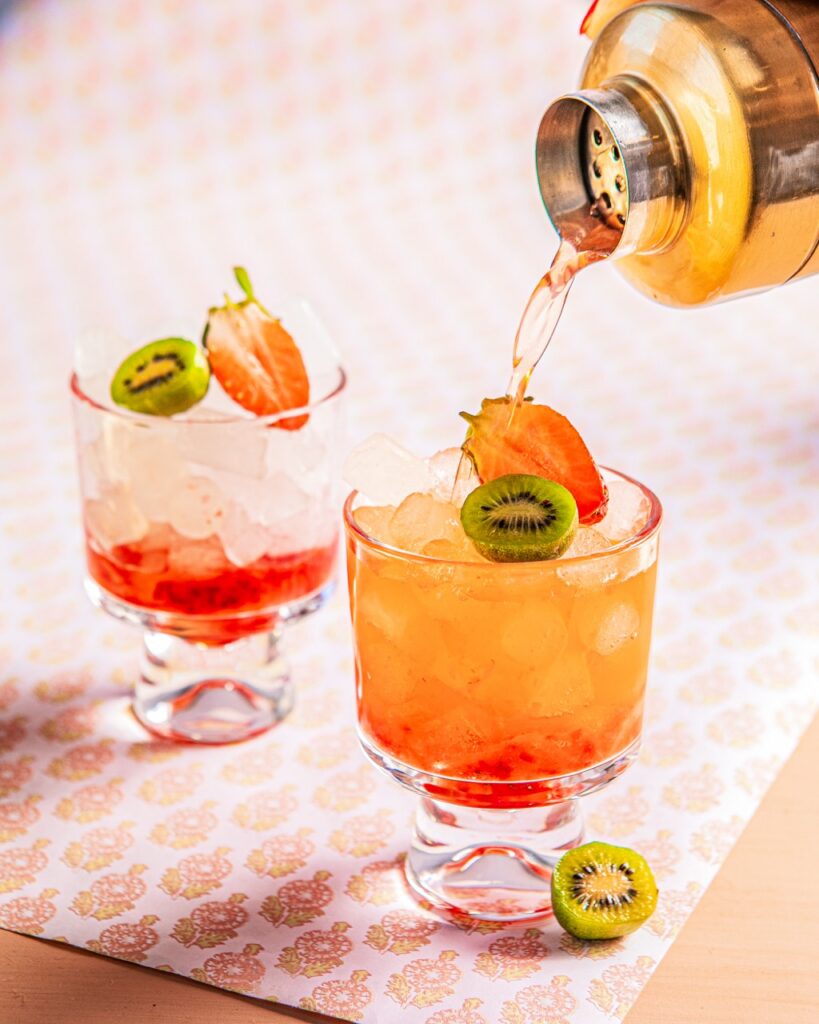 A cocktail pours from a cocktail shaker into a glass to make a Strawberry Kiwi Bramble with kiwi and strawberry garnish.