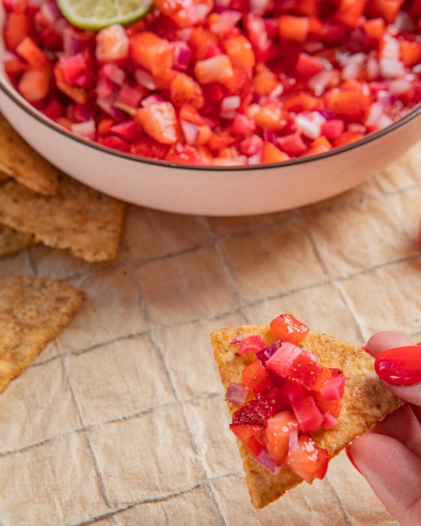 A person holds a tortilla chip with a strawberry salsa on it with a bowl at the top of the frame.
