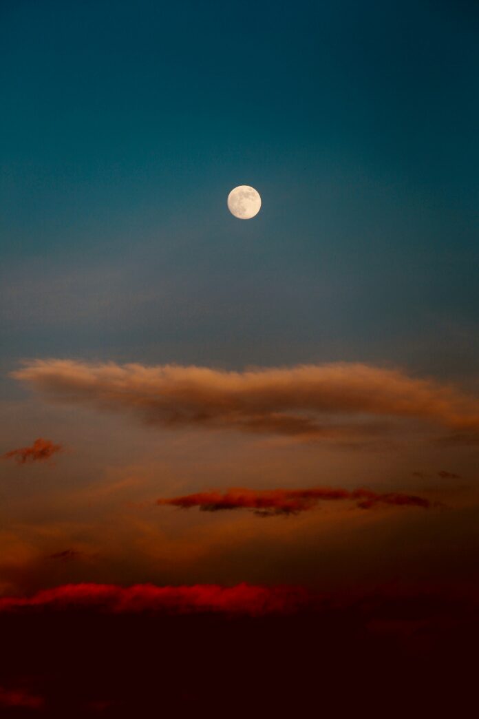 A full moon in a blue sky with orange-ish red clouds.