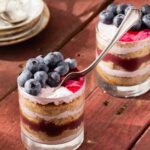 A spoon sticks out of a chai tea cake cup with blueberries on top in front of another chai tea cake cup with blueberries on top.