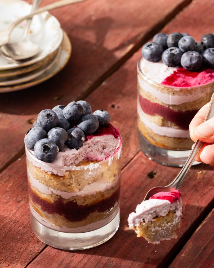 A person holds a spoon with a scoop of Chai Tea Cake with blueberry whipped cream beside two cups of the cake and frosting.