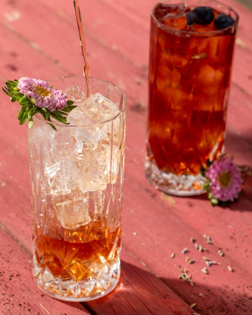 One glass fills with a tea-looking punch in a glass with ice and a flower garnish then another sits behind with dark punch and a blue berry garnish on a pink picnic table.