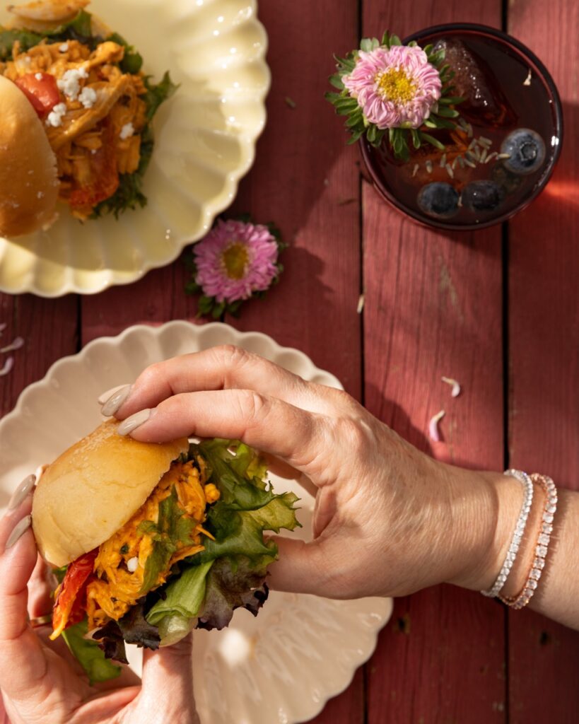 A woman holds a chicken slider with a bunch of toppings on top and a tea in the top right corner.