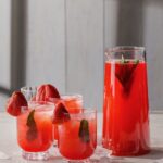 Three glasses of red punch beside a large pitcher of the punch with mint leaves inside and strawberries on the rim.
