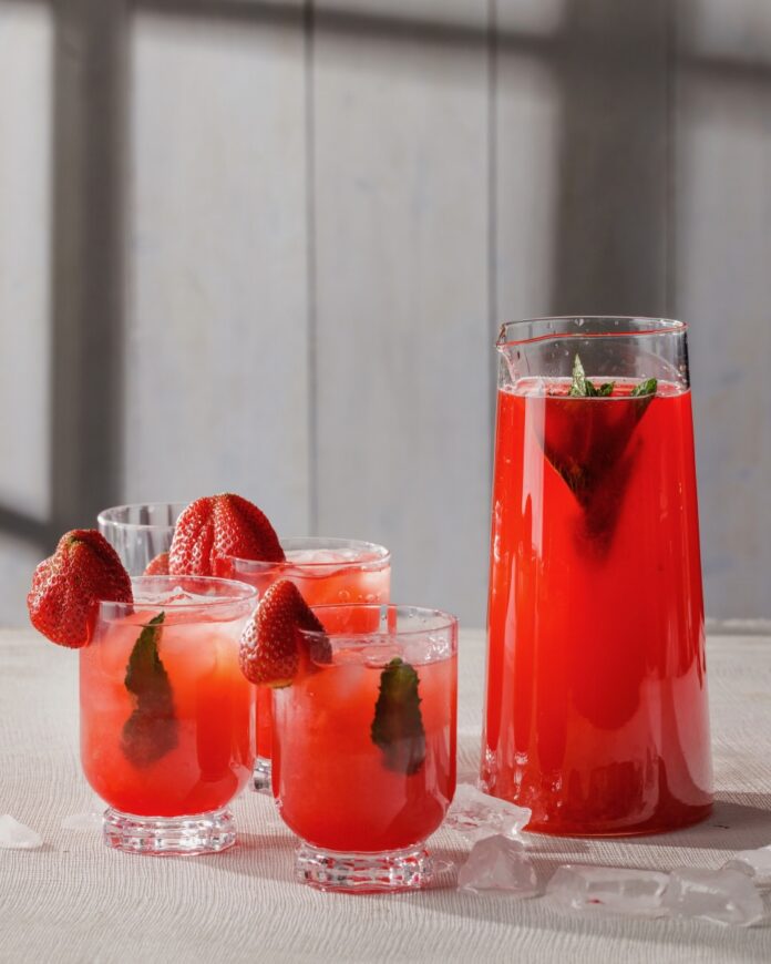Three glasses of red punch beside a large pitcher of the punch with mint leaves inside and strawberries on the rim.