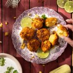 A person picks up a Mexican Street Cornbread fritter off a plate that sits beside slices limes, three forks, an ear of corn, and a bowl of lime crema with cilantro.