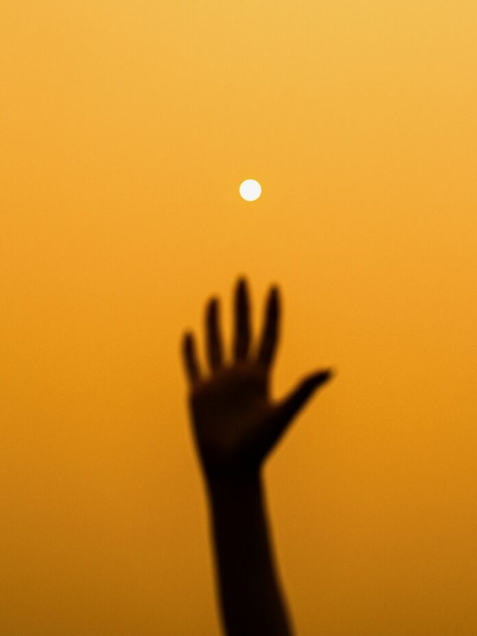 A person raises to hand up in a yellow/orange sky to touch the summer sun.