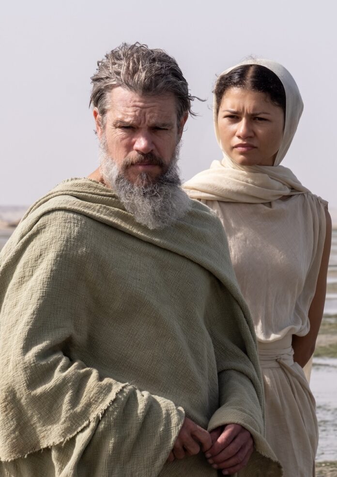 Matt Damon in The Odyssey stands in long cloths with a grey beard and hair as Zendaya stands behind him in long white cloths and scarves.