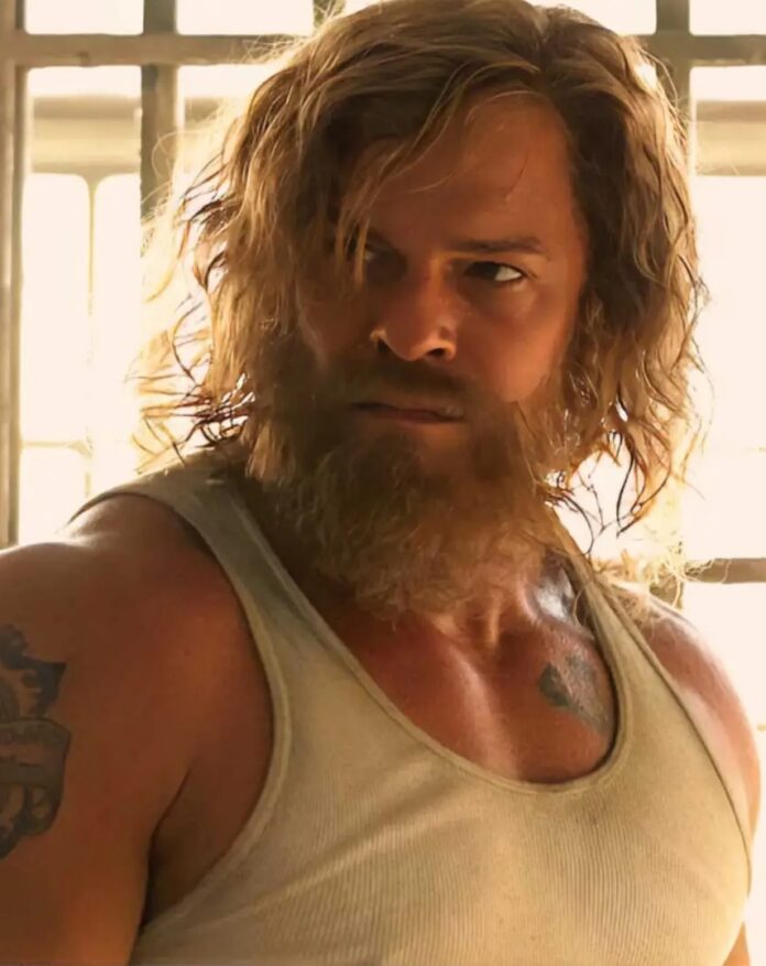 A man with long hair and a beard and muscles stands in a white muscle tee.