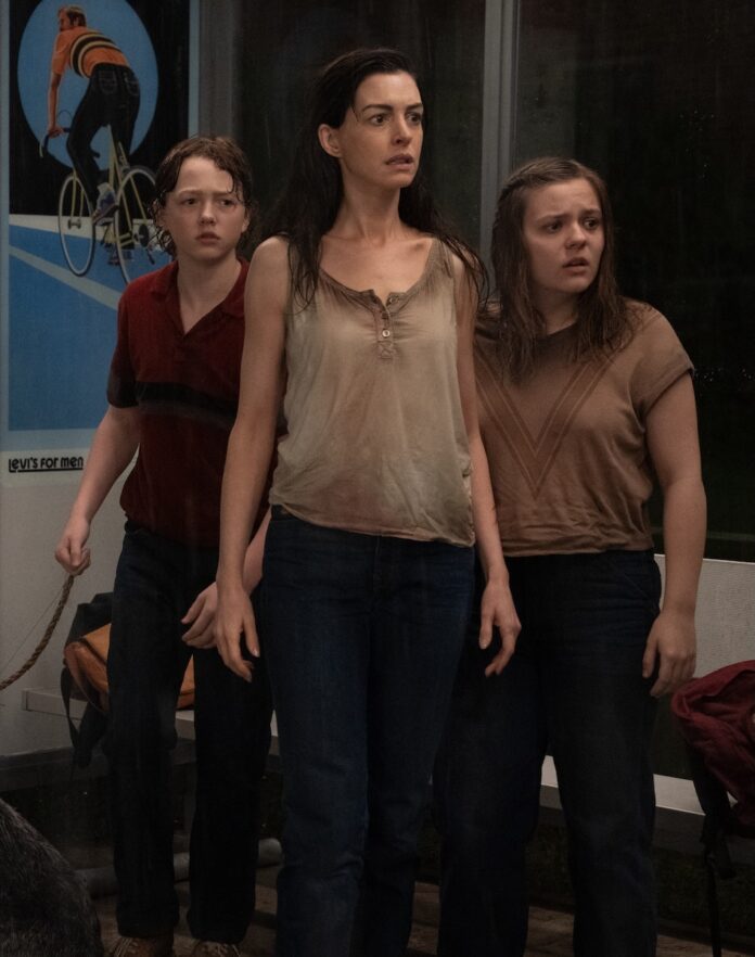 Anne Hathaway and two teens stand beside each other in dirty clothes.