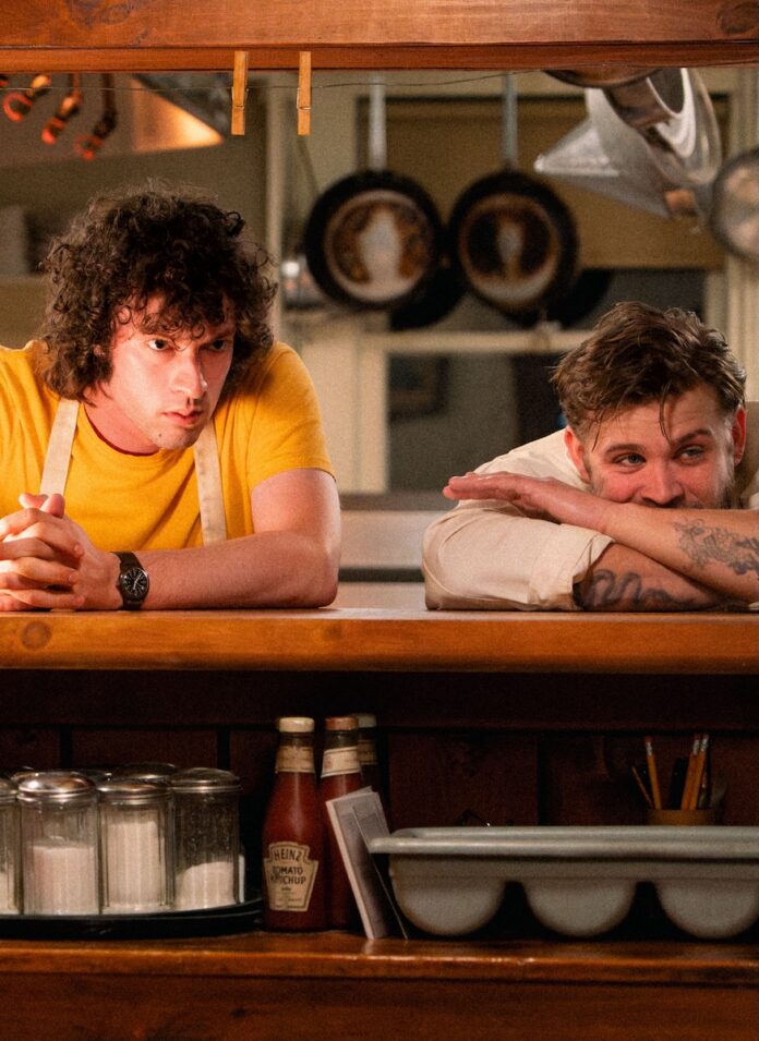 Dominic Sessa's character in Tony in a yellow shirt and apron behind a restaurant window with a Chef.