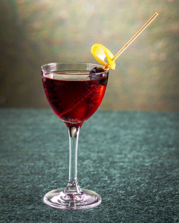 Artist in Residence cocktail in a stemmed glass, garnished with a Luxardo cherry wrapped in a lemon twist, featuring flavors of citrus gin, cherry, and chocolate bitters.