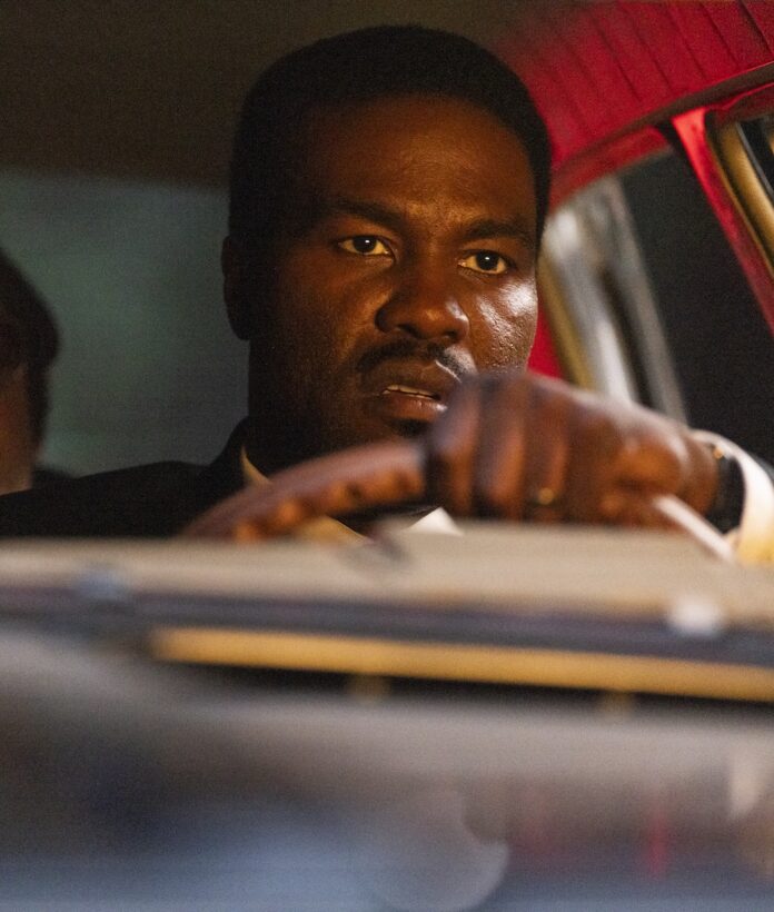 A man drives a car at night with red lights glinting off the inside of the car.