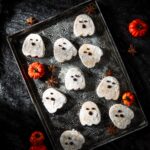 Ghost-shaped pumpkin brown butter shortbread cookies dusted with powdered sugar and decorated with melted chocolate faces, arranged on a vintage tray with mini pumpkins and star anise.