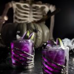 Poison Apple Punch in a two glasses topped with dry ice fog and purple steeped apple slices in front of a skeleton.