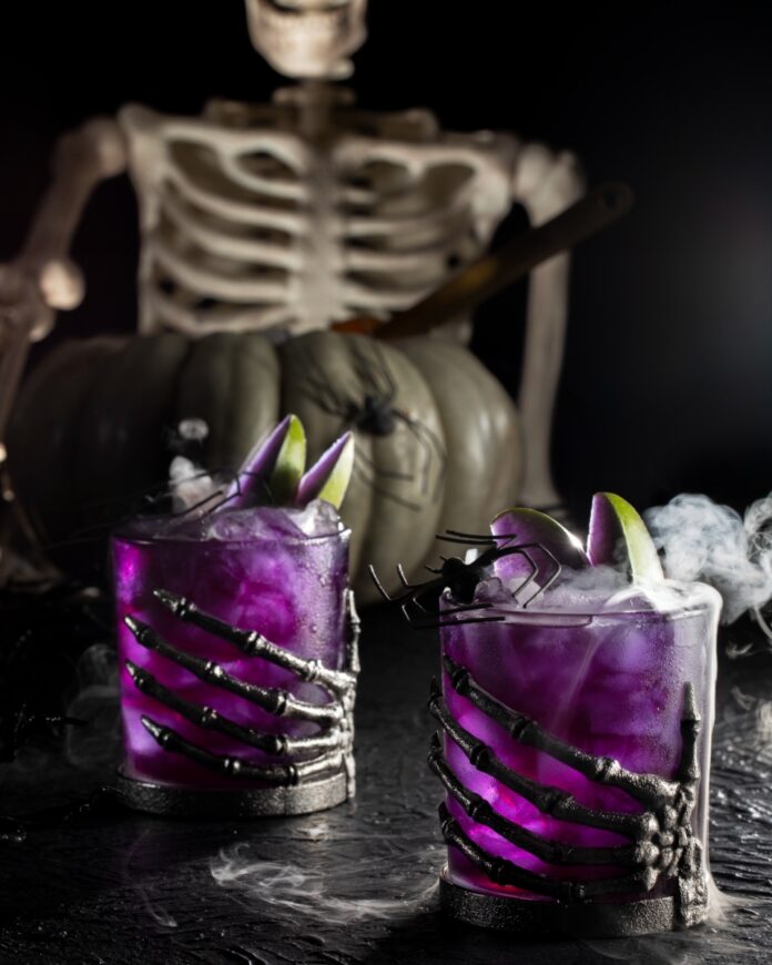 Poison Apple Punch Recipe A Magical Halloween Cocktail TABLE Magazine Poison Apple Punch in a two glasses topped with dry ice fog and purple steeped apple slices in front of a skeleton.