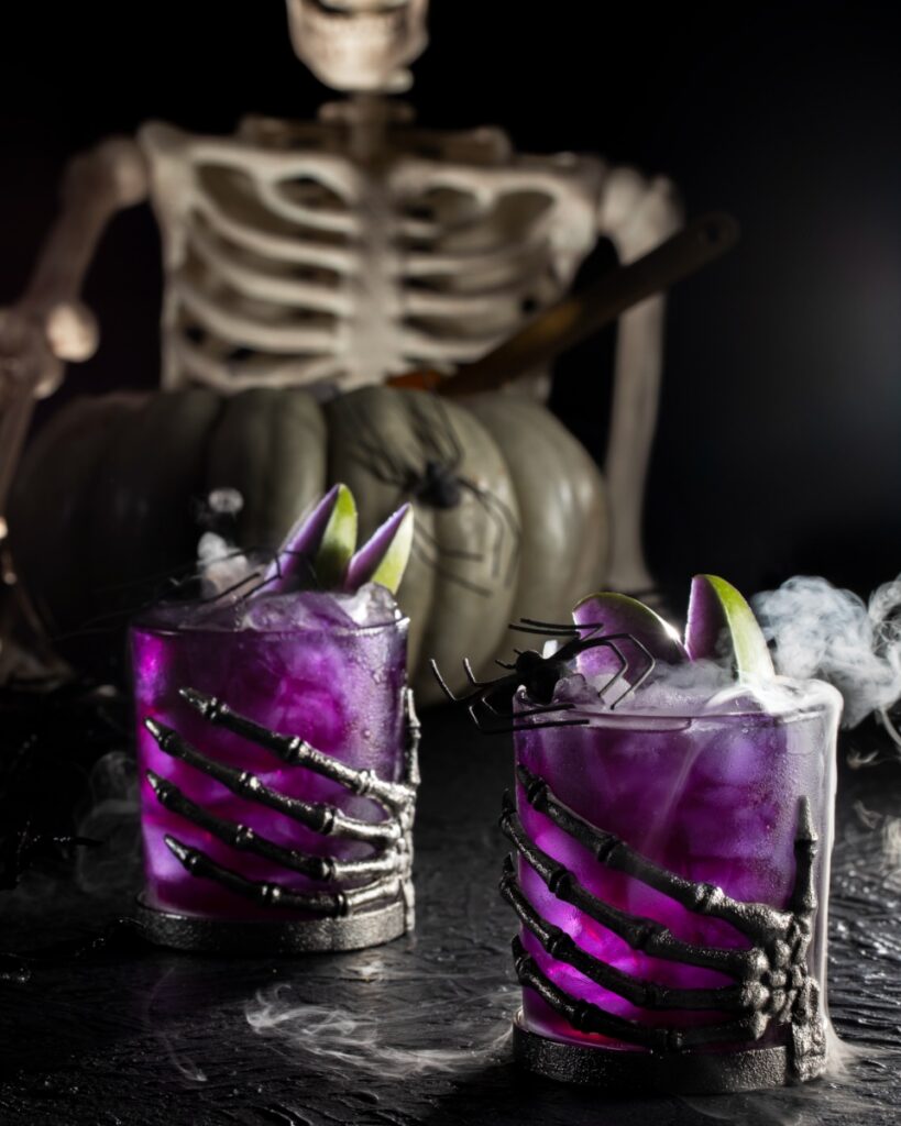 Poison Apple Punch in a two glasses topped with dry ice fog and purple steeped apple slices in front of a skeleton.