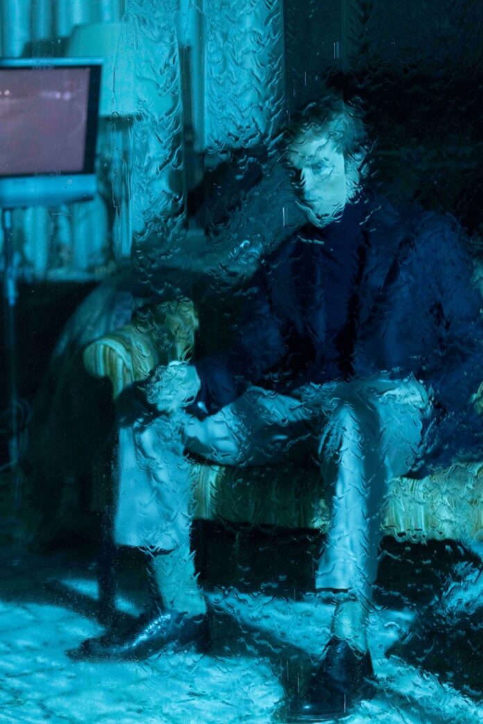 Primetime Featured TABLE Magazine Robert Pattinson in the movie Primetime sitting in blue light behind smeared glass.