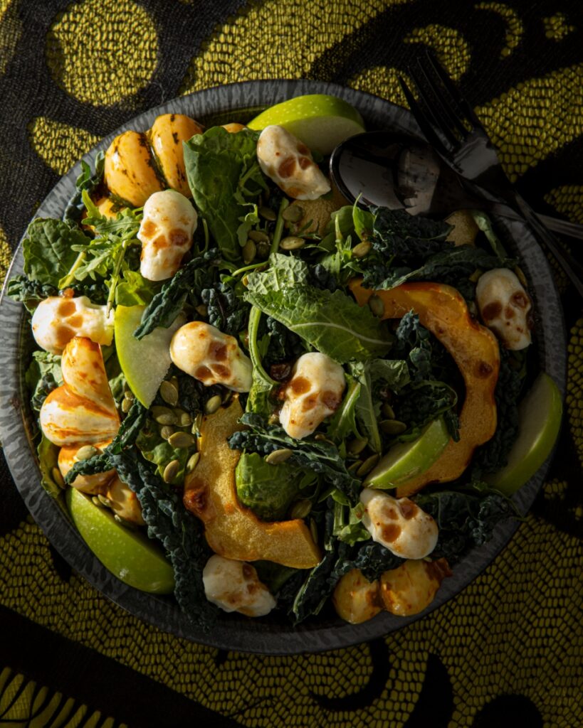 A serving bowl of roasted squash salad with kale, apple, and pepitas, topped with mozzarella skulls for Halloween.