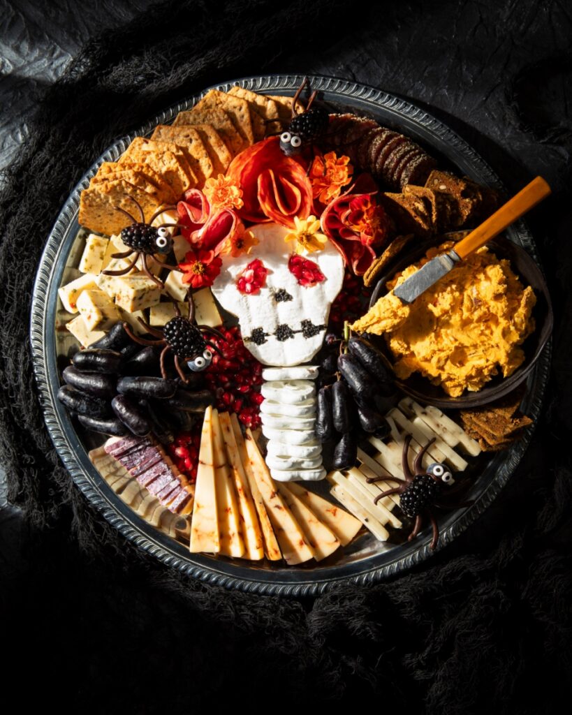 Halloween cheese board with a brie skull, salami wig, marigolds, blackberry spiders, black grapes, cheeses, crackers, and a bowl of savory pumpkin dip.