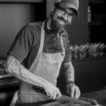 A black and white portrait of Chef Kevin Hermann cutting produce on a cutting board with an apron and hat on.