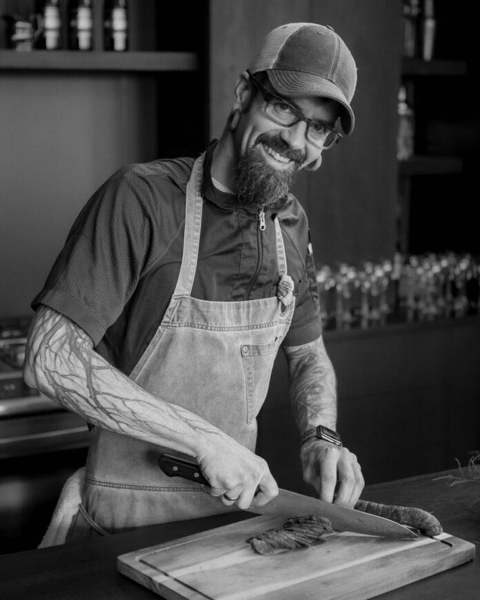A black and white portrait of Chef Kevin Hermann cutting produce on a cutting board with an apron and hat on.