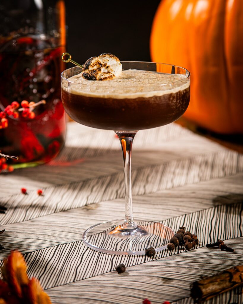 A pumpkin spice martini with a marshmallow garnish sits on a white table in front of a bottle of red liquor and a pumpkin.