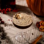 A pumpkin spice martini on a table with a marshmallow garnish sitting beside a pumpkin and bottle of red infused liquor.