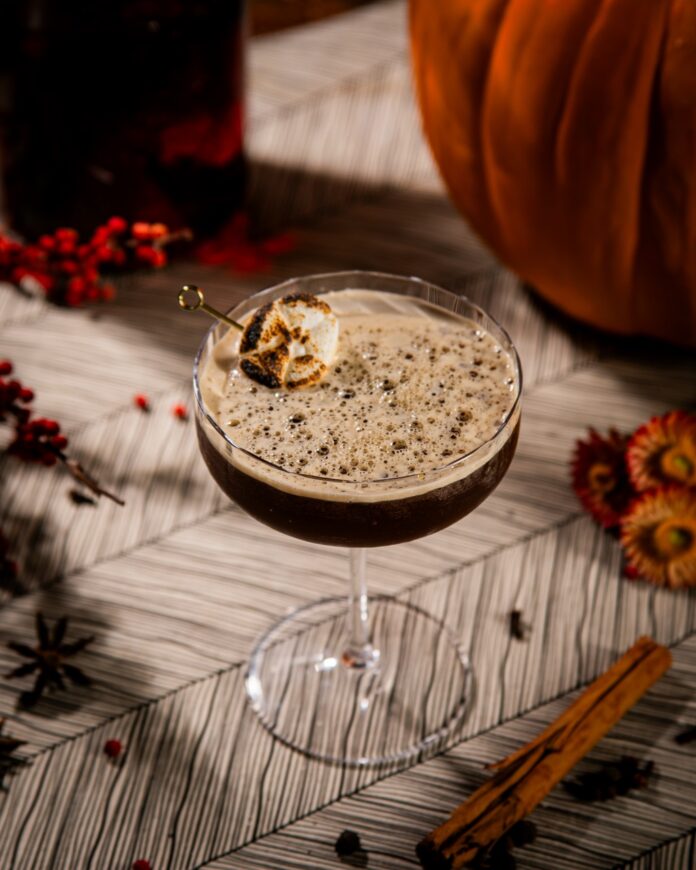 Pumpkin Spice Martini Recipe TABLE Magazine 3 A pumpkin spice martini on a table with a marshmallow garnish sitting beside a pumpkin and bottle of red infused liquor.