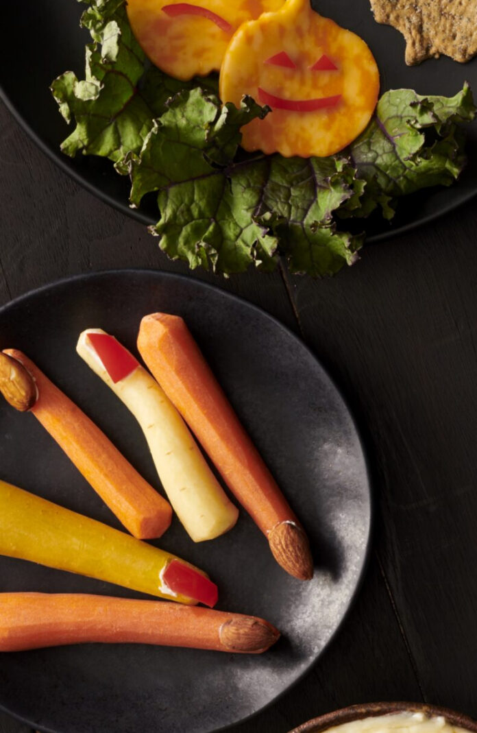 Carrot sticks on a black plate with red fingernails on them below two slices of cheese made to look like jack o lanterns.