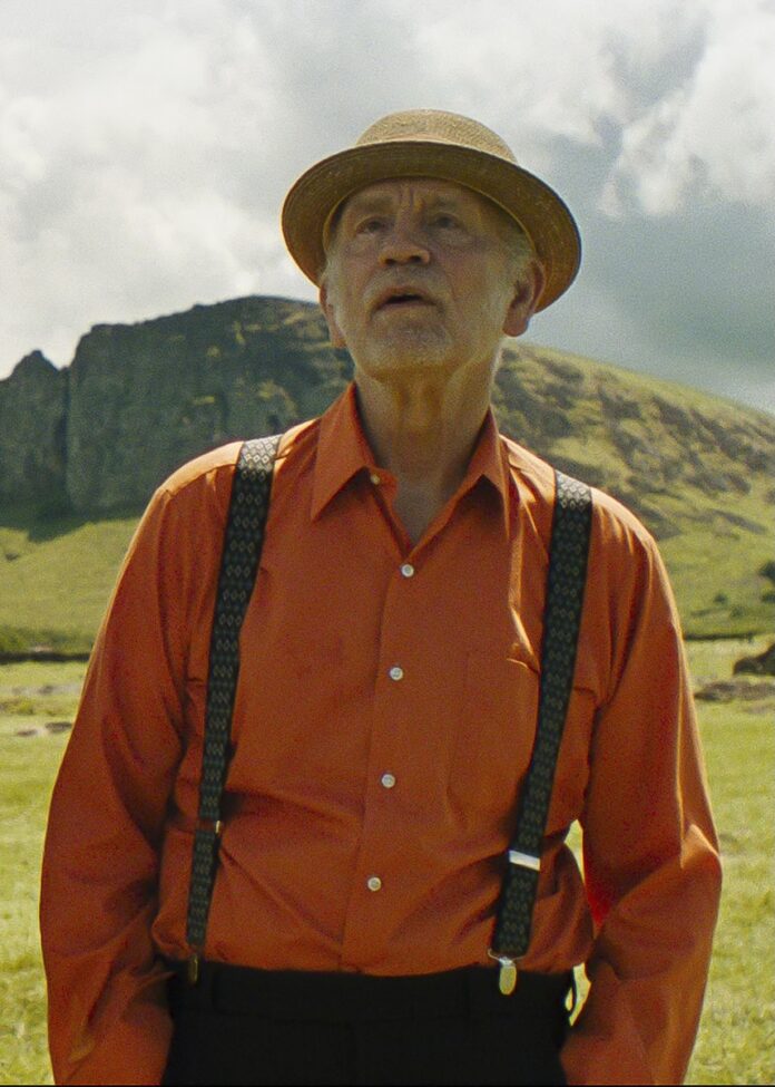 A man stands in an orange button down shirt with black suspenders and a brown hat in Wild Horse Nine.