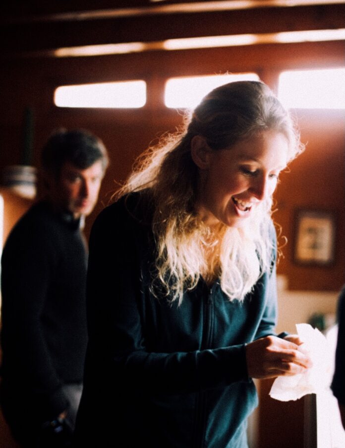 Elizabeth Holmes stands in her kitchen with Nathan Fielder behind her in the background.